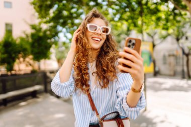 Güneş gözlüğü takan ve telefon kullanan gülümseyen kadın güneşli bir günde bankta oturur. Telefonunu kullanan genç bir kadın parkta yürümekten zevk alıyor ve bankta dinleniyor. İletişim ve rahatlama kavramları.