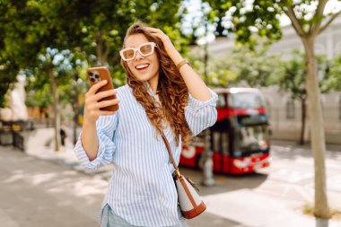 Güneş gözlüğü takan ve telefon kullanan gülümseyen kadın güneşli bir günde bankta oturur. Telefonunu kullanan genç bir kadın parkta yürümekten zevk alıyor ve bankta dinleniyor. İletişim ve rahatlama kavramları.