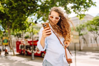 Güneş gözlüğü takan ve telefon kullanan gülümseyen kadın güneşli bir günde bankta oturur. Telefonunu kullanan genç bir kadın parkta yürümekten zevk alıyor ve bankta dinleniyor. İletişim ve rahatlama kavramları.