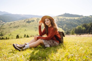 Güneşli bir günde yeşil bir tepede sırt çantası ve şapkası olan genç bir kadın. Gezgin, dağların özgürlüğünü hissederek dağ manzarasının tadını çıkarıyor. Macera ve rahatlama kavramları.