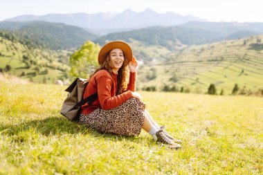 Güneşli bir günde yeşil bir tepede sırt çantası ve şapkası olan genç bir kadın. Gezgin, dağların özgürlüğünü hissederek dağ manzarasının tadını çıkarıyor. Macera ve rahatlama kavramları.