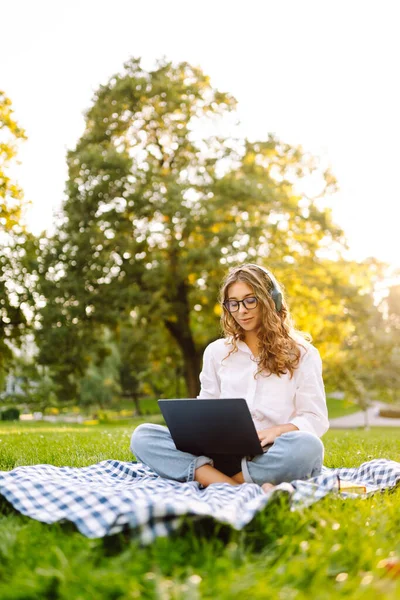 Sunny Park 'ta elinde dizüstü bilgisayarla battaniyenin üzerinde oturan serbest çalışan bir kadın. Kablosuz kulaklık takan kadın laptopta çalışıyor ve açık havada güneşli bir günün tadını çıkarıyor. Kavram, teknoloji, doğa ve rahatlama..