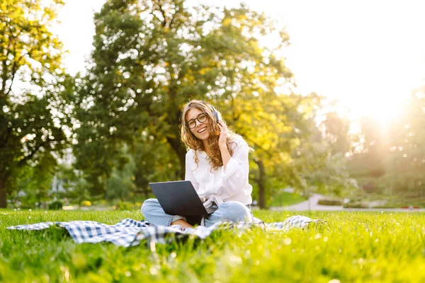 Sunny Park 'ta elinde dizüstü bilgisayarla battaniyenin üzerinde oturan serbest çalışan bir kadın. Kablosuz kulaklık takan kadın laptopta çalışıyor ve açık havada güneşli bir günün tadını çıkarıyor. Kavram, teknoloji, doğa ve rahatlama..