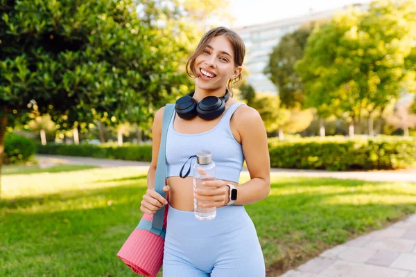 Kablosuz kulaklıkları ve yoga minderiyle atletik kadın güneşli parkta su içiyor. Genç bir kadın idmandan sonra susuzluğunu giderir ve gün batımında müzik dinlemekten hoşlanır. Spor ve rahatlama kavramı.