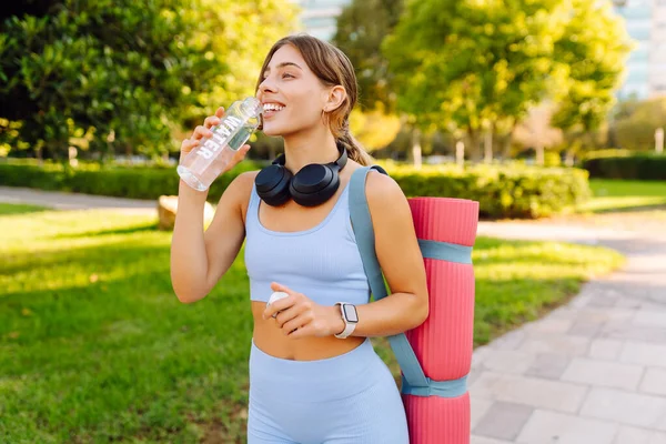 Kablosuz kulaklıkları ve yoga minderiyle atletik kadın güneşli parkta su içiyor. Genç bir kadın idmandan sonra susuzluğunu giderir ve gün batımında müzik dinlemekten hoşlanır. Spor ve rahatlama kavramı.