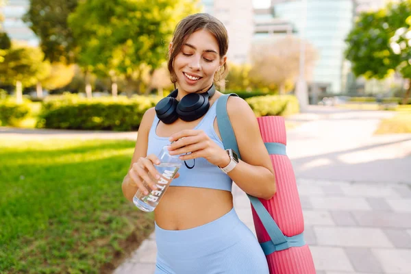 Kablosuz kulaklıkları ve yoga minderiyle atletik kadın güneşli parkta su içiyor. Genç bir kadın idmandan sonra susuzluğunu giderir ve gün batımında müzik dinlemekten hoşlanır. Spor ve rahatlama kavramı.