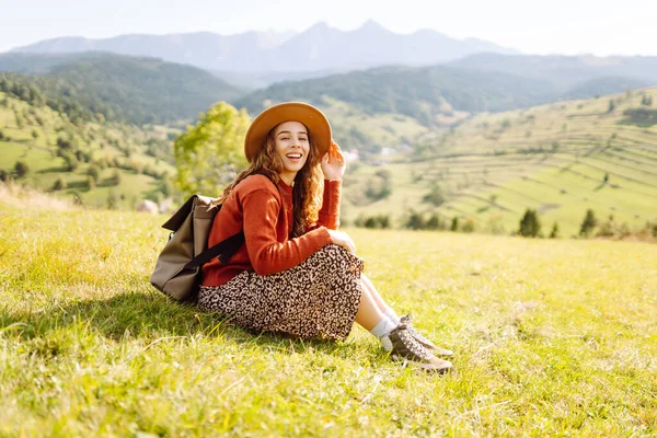 Güneşli bir günde yeşil bir tepede sırt çantası ve şapkası olan genç bir kadın. Gezgin, dağların özgürlüğünü hissederek dağ manzarasının tadını çıkarıyor. Macera ve rahatlama kavramları.