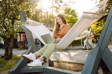 Bilgisayarlı neşeli kadın parkta hamakta oturuyor. Genç kadın hamakta yatarken dizüstü bilgisayarda çalışıyor ve gün batımında doğada dinlenmekten hoşlanıyor. Serbest çalışma, gevşeme, üretkenlik kavramları.