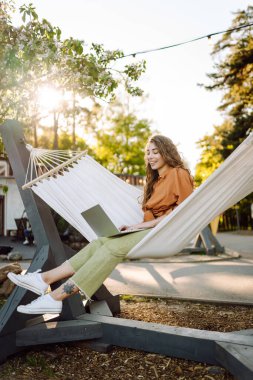 Bilgisayarlı neşeli kadın parkta hamakta oturuyor. Genç kadın hamakta yatarken dizüstü bilgisayarda çalışıyor ve gün batımında doğada dinlenmekten hoşlanıyor. Serbest çalışma, gevşeme, üretkenlik kavramları.