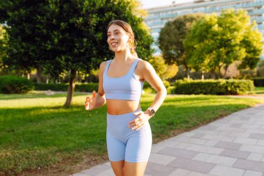 Güneşli bir parkta koşan gülümseyen bir kadın. Atletik bir kadın patika boyunca koşar ve güzel havanın tadını çıkarır. Koşmak, spor yapmak ve sağlıklı bir yaşam tarzı..