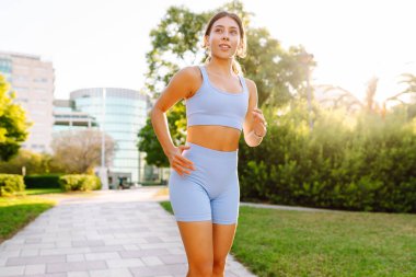 Güneşli bir parkta koşan gülümseyen bir kadın. Atletik bir kadın patika boyunca koşar ve güzel havanın tadını çıkarır. Koşmak, spor yapmak ve sağlıklı bir yaşam tarzı..