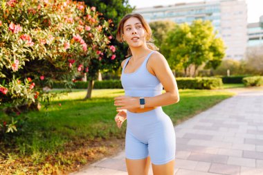 Güneşli bir parkta koşan gülümseyen bir kadın. Atletik bir kadın patika boyunca koşar ve güzel havanın tadını çıkarır. Koşmak, spor yapmak ve sağlıklı bir yaşam tarzı..