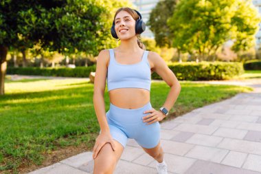 Spor kıyafetli bir kadın antrenmandan önce esniyor, kablosuz kulaklık takıyor, güneşli bir parkta. Atletik kadın egzersiz yapıyor, müzik dinlemekten hoşlanıyor. Spor, güzellik ve sağlık kavramı.