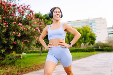 Spor kıyafetli bir kadın antrenmandan önce esniyor, kablosuz kulaklık takıyor, güneşli bir parkta. Atletik kadın egzersiz yapıyor, müzik dinlemekten hoşlanıyor. Spor, güzellik ve sağlık kavramı.