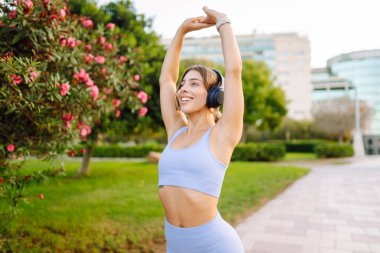 Spor kıyafetli bir kadın antrenmandan önce esniyor, kablosuz kulaklık takıyor, güneşli bir parkta. Atletik kadın egzersiz yapıyor, müzik dinlemekten hoşlanıyor. Spor, güzellik ve sağlık kavramı.
