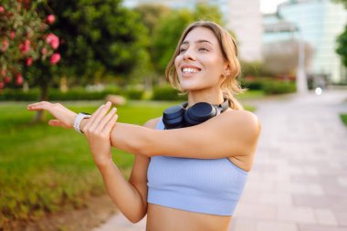 Spor kıyafetli bir kadın antrenmandan önce esniyor, kablosuz kulaklık takıyor, güneşli bir parkta. Atletik kadın egzersiz yapıyor, müzik dinlemekten hoşlanıyor. Spor, güzellik ve sağlık kavramı.