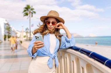 Şapka takan ve elinde telefon tutan genç bir kadın güneşli şehir seti boyunca yürüyor. Sırt çantalı güzel bir turist açık havada güneşli bir günün tadını çıkarıyor. Turizm konsepti, mesaj. Yaşam biçimi.