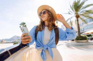 Şapka takan ve elinde telefon tutan genç bir kadın güneşli şehir seti boyunca yürüyor. Sırt çantalı güzel bir turist açık havada güneşli bir günün tadını çıkarıyor. Turizm konsepti, mesaj. Yaşam biçimi.