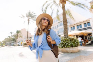 Şapka takan ve elinde telefon tutan genç bir kadın güneşli şehir seti boyunca yürüyor. Sırt çantalı güzel bir turist açık havada güneşli bir günün tadını çıkarıyor. Turizm konsepti, mesaj. Yaşam biçimi.