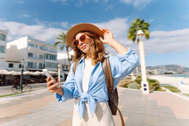 Şapka takan ve elinde telefon tutan genç bir kadın güneşli şehir seti boyunca yürüyor. Sırt çantalı güzel bir turist açık havada güneşli bir günün tadını çıkarıyor. Turizm konsepti, mesaj. Yaşam biçimi.