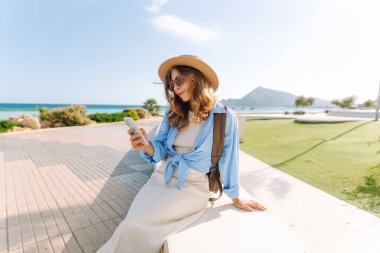 Şapka takan ve elinde telefon tutan genç bir kadın güneşli şehir seti boyunca yürüyor. Sırt çantalı güzel bir turist açık havada güneşli bir günün tadını çıkarıyor. Turizm konsepti, mesaj. Yaşam biçimi.