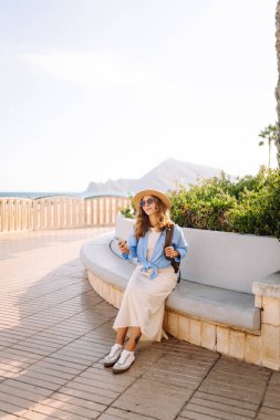 Şapka takan ve elinde telefon tutan genç bir kadın güneşli şehir seti boyunca yürüyor. Sırt çantalı güzel bir turist açık havada güneşli bir günün tadını çıkarıyor. Turizm konsepti, mesaj. Yaşam biçimi.