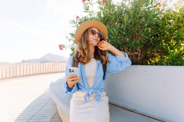 Şapka takan ve elinde telefon tutan genç bir kadın güneşli şehir seti boyunca yürüyor. Sırt çantalı güzel bir turist açık havada güneşli bir günün tadını çıkarıyor. Turizm konsepti, mesaj. Yaşam biçimi.
