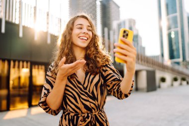 Bir kadın blogcu güneşli bir caddede telefonundan yayın yapıyor. Güzel bir kadın telefonuyla gezer ve güneşli günün tadını çıkarır. Blog konsepti, video çağrısı, şehir. Yaşam biçimi.