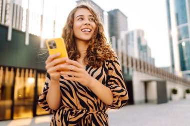 Bir kadın blogcu güneşli bir caddede telefonundan yayın yapıyor. Güzel bir kadın telefonuyla gezer ve güneşli günün tadını çıkarır. Blog konsepti, video çağrısı, şehir. Yaşam biçimi.