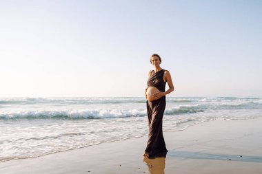 Hamile bir kadın güneşin altında kumlu bir plajda yürüyor. Bir çocuk bekleyen genç bir kadın gün batımında deniz manzarasının tadını çıkarıyor, karnına dokunuyor. Doğa, keyif ve rahatlama kavramları.