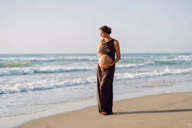 Hamile bir kadın güneşin altında kumlu bir plajda yürüyor. Bir çocuk bekleyen genç bir kadın gün batımında deniz manzarasının tadını çıkarıyor, karnına dokunuyor. Doğa, keyif ve rahatlama kavramları.