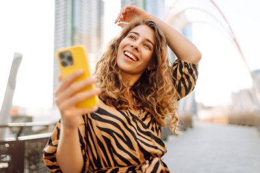 Parlak bir telefon tutan genç bir kadının portresi güneşli bir günde şehir manzarasının tadını çıkarıyor. Güzel bir kadın dışarıda blog yazıyor. Konseptler: selfie, kentsel, turizm.