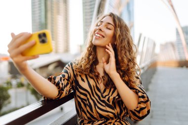 Parlak bir telefon tutan genç bir kadının portresi güneşli bir günde şehir manzarasının tadını çıkarıyor. Güzel bir kadın dışarıda blog yazıyor. Konseptler: selfie, kentsel, turizm.