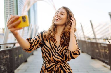 Parlak bir telefon tutan genç bir kadının portresi güneşli bir günde şehir manzarasının tadını çıkarıyor. Güzel bir kadın dışarıda blog yazıyor. Konseptler: selfie, kentsel, turizm.