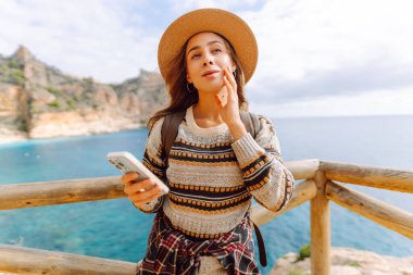 Şapka takan ve sırt çantası taşıyan kadın gezgin, gün batımında telefonuyla izleme platformunda duruyor. Telefonu olan gülümseyen kadın uçurumda deniz manzarasını seviyor. Konseptler: macera, teknoloji.