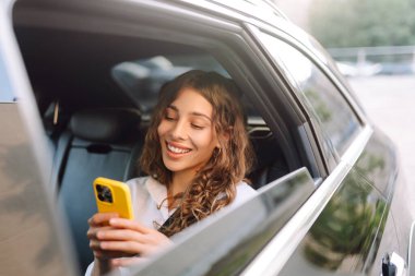 Bir arabanın arka koltuğunda oturan, elinde telefon tutan genç bir kadının portresi. Güneşli bir günde güzel bir kadın takside telefonuna mesaj atıyor. Ulaşım, teknoloji ve şehir kavramları.