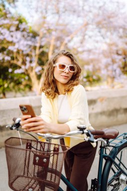 Güneşli bir parkta telefonu olan gülümseyen bir kadın bisikletinin yanında duruyor. Genç bir turist bisiklet sürüyor ve telefonuyla güneşli bir günün tadını çıkarıyor. Kavram: Hafta sonu, eğlence, aktif yaşam tarzı.