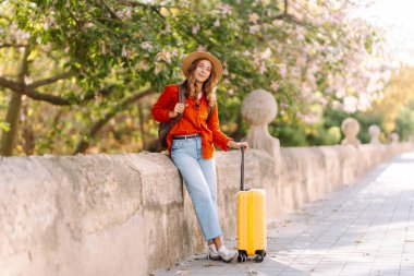 Parlak renkli bavulu olan neşeli bir kadın güneşli bir şehir caddesinde geziniyor. Şapkalı ve sırt çantalı genç bir turist güneşli günün tadını çıkarıyor. Kavram: Turizm, eğlence ve macera.