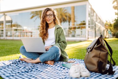 Kadın bir serbest çalışan güneşli bir parkta minder üzerinde otururken dizüstü bilgisayarda çalışır. Gözlüklü genç bir kadın dizüstü bilgisayarla uzaktan çalışmayı ve gün batımında dinlenmeyi sever. Serbest meslek ve hafta sonu kavramları.