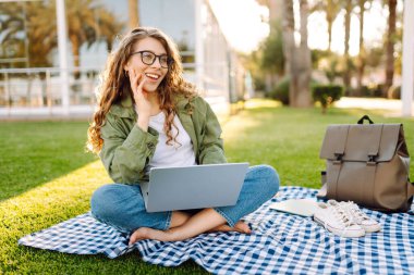 Kadın bir serbest çalışan güneşli bir parkta minder üzerinde otururken dizüstü bilgisayarda çalışır. Gözlüklü genç bir kadın dizüstü bilgisayarla uzaktan çalışmayı ve gün batımında dinlenmeyi sever. Serbest meslek ve hafta sonu kavramları.