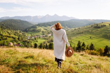 Güneşli bir günde yeşil bir tepe boyunca yürüyen şapkalı genç bir kadın. Arkadan bak. Güzel kadın kendini özgür hissediyor ve gün batımında dağ manzarasının tadını çıkarıyor. Doğa ve özgürlük kavramı.