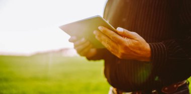 Bir adam yeşil bir alanda duruyor, akıllı telefonuna odaklanmış. Güneş arkasından batıyor, o cihazla temas kurarken vücuduna ışık tutuyor. Kırsal alanda akşam vaktidir..