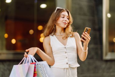 Güneşli bir günde genç bir kadın alışveriş torbaları ve telefonuyla şehirde duruyor. Alışverişten sonra güzel bir kadın, yürüyüşten ve telefonundan mesaj atmaktan zevk alıyor. Tüketim kavramı, alışveriş.