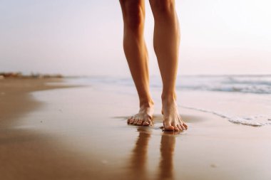 Genç bir kadının, batan güneşin ışınlarıyla kumda suyla yürüyen bacaklarının yakın çekimi. Kadın temiz havada deniz kıyısında yürümekten zevk alıyor. Konsept: yürüyüş, turizm, özgürlük.