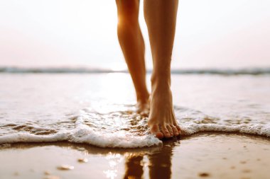 Genç bir kadının, batan güneşin ışınlarıyla kumda suyla yürüyen bacaklarının yakın çekimi. Kadın temiz havada deniz kıyısında yürümekten zevk alıyor. Konsept: yürüyüş, turizm, özgürlük.