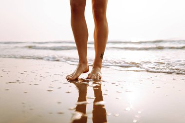 Genç bir kadının, batan güneşin ışınlarıyla kumda suyla yürüyen bacaklarının yakın çekimi. Kadın temiz havada deniz kıyısında yürümekten zevk alıyor. Konsept: yürüyüş, turizm, özgürlük.