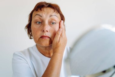 Güneşli odada aynada yansımayı inceleyen olgun bir kadının portresi. Güzel bir kadın, rahat bir odada yaşlanma noktalarını ve kırışıklıkları inceler. Güzellik, doğallık ve rahatlama kavramı.