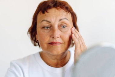 Güneşli odada aynada yansımayı inceleyen olgun bir kadının portresi. Güzel bir kadın, rahat bir odada yaşlanma noktalarını ve kırışıklıkları inceler. Güzellik, doğallık ve rahatlama kavramı.