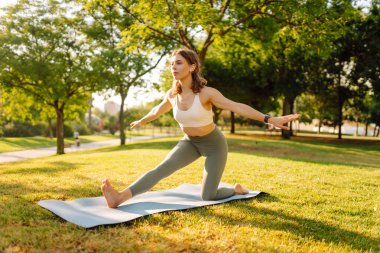Atletik kıyafetleri ve kablosuz kulaklıkları olan güzel bir kadın parkta yoga yapıyor. Atletik kadın güneşli bir günün tadını çıkarıyor, meditasyon yapıyor ya da dışarıda egzersiz yapıyor. Egzersiz ve rahatlama kavramları.