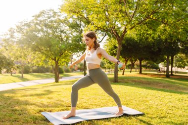 Atletik kıyafetleri ve kablosuz kulaklıkları olan güzel bir kadın parkta yoga yapıyor. Atletik kadın güneşli bir günün tadını çıkarıyor, meditasyon yapıyor ya da dışarıda egzersiz yapıyor. Egzersiz ve rahatlama kavramları.